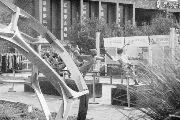 Ilford HP5 Plus 400 (FF)

English Alt Text: An outdoor art installation features dynamic sculptures of children in playful poses, appearing to run or fly with joyful expressions. Each child sculpture is mounted on a separate platform and arranged in a line, creating a sense of motion and interaction. In the foreground, a large abstract metal structure frames the scene. Behind the sculptures are display boards with text and images, likely describing the exhibit. A building with vertical banners …