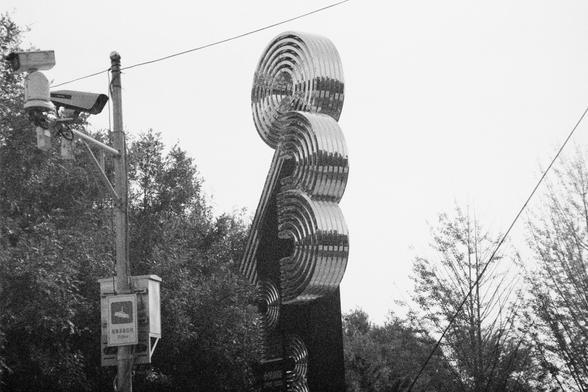Ilford HP5 Plus 400 (FF)

English Alt Text:  
A black and white photo of a modern urban scene featuring a large abstract metallic sculpture and surveillance equipment. The sculpture consists of stacked concentric metal rings curving upward in a spiral, resembling a futuristic tower. It stands among trees, suggesting a public park or plaza. To the left, a utility pole supports two surveillance cameras and a box with signage. The box displays Chinese characters and the word “Video,” along with a …