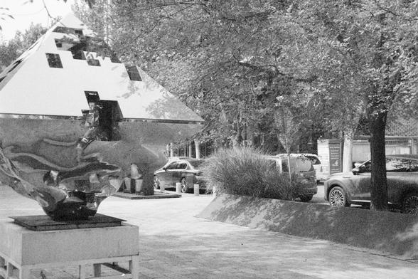 Ilford HP5 Plus 400 (FF)

English Alt Text:  
An outdoor urban scene with a striking abstract sculpture as the focal point. The sculpture has a mirrored surface and a geometric design: a pyramid-like top transitions into a fluid, distorted base. It sits on a white pedestal in a landscaped plaza with ornamental grasses and trees. Parked cars line the background street, and a signboard is visible nearby. The sculpture’s reflective surface captures surrounding light and movement, creating a dynami…