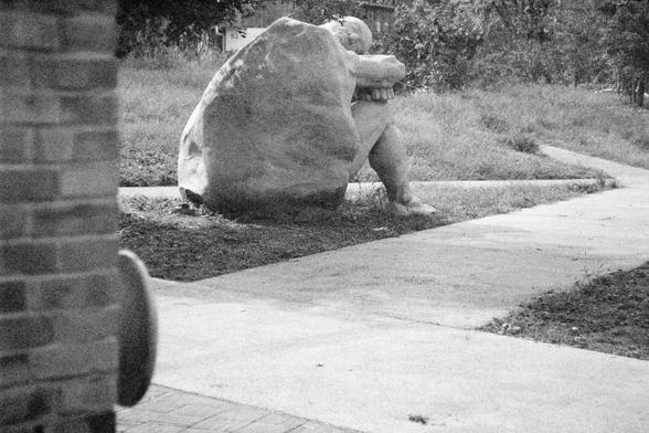 Ilford HP5 Plus 400 (FF)

English Alt Text:  
A stone sculpture of a person seated on the ground with knees drawn up, arms resting on the knees, and head bowed. The figure appears to be emerging from or partially enveloped by a rough-textured stone, blending human form with natural material. Set on a grassy area beside a sidewalk, the sculpture is surrounded by trees and a distant building, suggesting a park or campus. The posture conveys introspection, solitude, or emotional resilience, inviti…