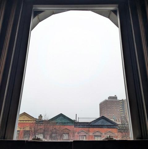 Looking through an arched window seventy-six minutes after sunrise the sky is a dense white mass. Pointed roofs of Harlem brownstones with red brickwork are across the street, and a taller apartment building can be seen in the distance. The leafless tops of two trees are on the bottom and right.