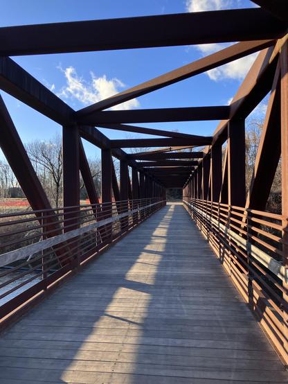 An empty pedestrian bridge featuring metal framework and wooden flooring. Bright blue sky with a few clouds visible above. Shadows cast across the bridge surface.