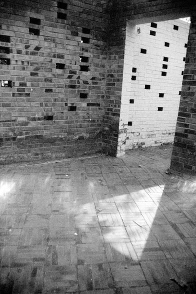 ERA 100 (FF)

English Alt Text:
A black-and-white photo of an interior brick corner with three walls. The left and right walls are made of dark bricks, while the center wall is white. Each wall has a pattern of missing bricks, forming small rectangular openings. Sunlight streams through these gaps, casting geometric shadows on the tiled floor. A prominent triangle of light appears on the floor, emphasizing the interplay between architecture and natural light. The image evokes a sense of quiet a…