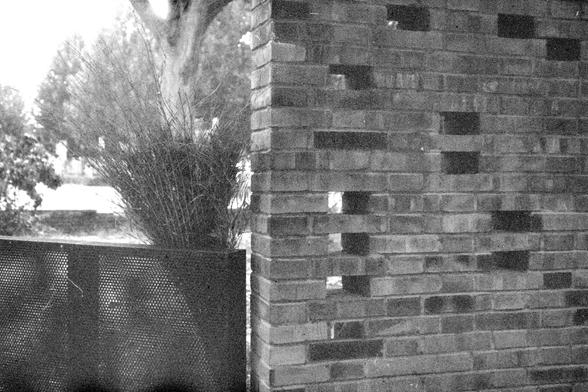 ERA 100 (FF)

English Alt Text:
A black-and-white image showing a section of a brick wall with several bricks intentionally left out, creating small rectangular openings. To the left, a perforated metal panel stands beside a broomstick. In the background, trees and bright daylight are visible. The design of the wall blends solid structure with open space, possibly for ventilation or aesthetic effect. The image balances nature and architecture, with light filtering through the gaps to create a s…