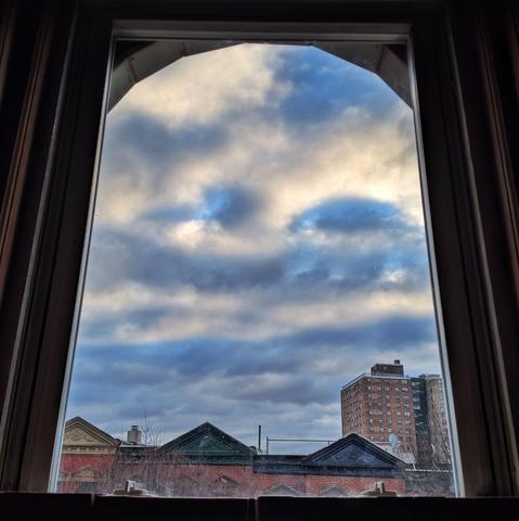 Looking through an arched window forty-five minutes after sunrise the sky is filled with lumpy gray and white clouds with patches of blue showing through. Pointed roofs of Harlem brownstones with red brickwork are across the street, and a taller apartment building can be seen in the distance. The leafless tops of two trees are on the bottom and right.