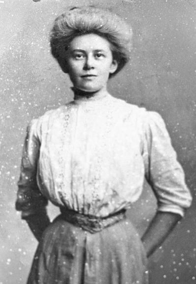 Photo of a photograph of a white woman standing square to the camera, wearing a white lace blouse and a light-colored skirt.