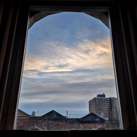 Looking through an arched window fourteen minutes after sunrise the blue sky is filled with diffuse horizontal streaks of and gray clouds. Pointed roofs of Harlem brownstones with red brickwork are across the street, and a taller apartment building can be seen in the distance. The leafless tops of two trees are on the bottom and right. The window is grimy on the bottom.