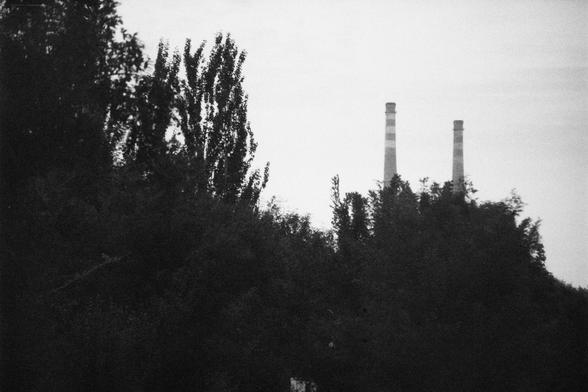 ERA 100 (FF)

English Alt Text:
A black-and-white photograph shows a dense forest in the foreground with two tall industrial smokestacks rising in the background. The smokestacks have horizontal bands and are partially obscured by treetops. The sky is overcast, casting a muted light over the scene. The contrast between the natural greenery and the industrial structures evokes themes of environmental tension and coexistence.

中文替代文字：
这是一张黑白照片，前景是茂密的森林，背景中有两根高大的工业烟囱从树顶后方升起。烟囱上有水平条纹，部分被树冠遮挡。天空阴沉，光…