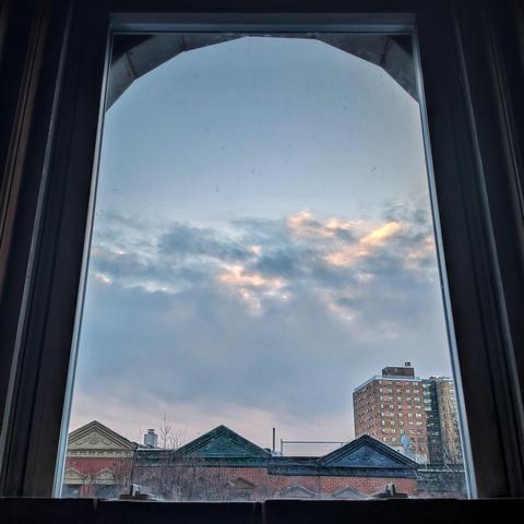 Looking through an arched window twenty minutes after sunrise a large storm bank is moving East away from us, leaving the upper sky clear and blue and the lower half of the sky full of turbulent white and gray clouds. Pointed roofs of Harlem brownstones with red brickwork are across the street, and a taller apartment building can be seen in the distance. The leafless tops of two trees are on the bottom and right.