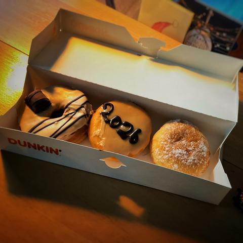Drei Pfannkuchen aka „Donuts“ in einer Schachtel mit der Aufschrift „Dunkin‘“. Der linke mit Zuckerguss, Schoko-Drizzle und einem Kinder Bueno, der mittlere trägt auf dem Zuckerguss den Schriftzug „2026“ in Schokolade geschrieben, der rechte ist mit Puderzucker bestäubt.