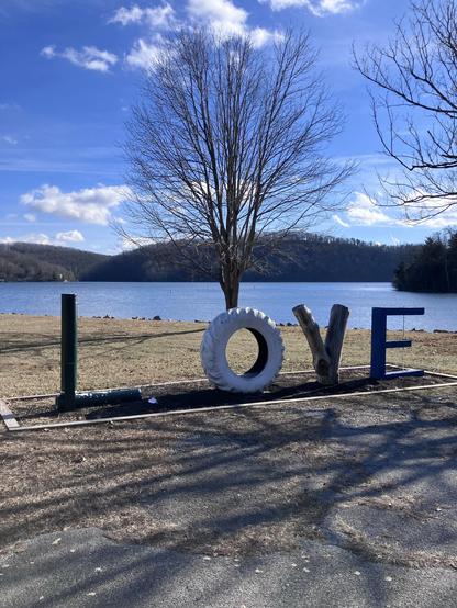 A serene lakeside scene featuring a large sign spelling "LOVE" with the letters made of various materials, including a white tire and wood. In the background, there is a calm lake and a few trees against a blue sky with scattered clouds.