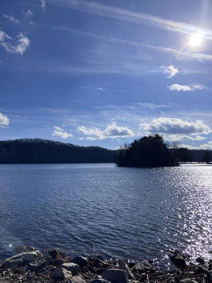 A serene lakeside scene with calm water reflecting sunlight, surrounded by trees and hills under a blue sky with scattered clouds.