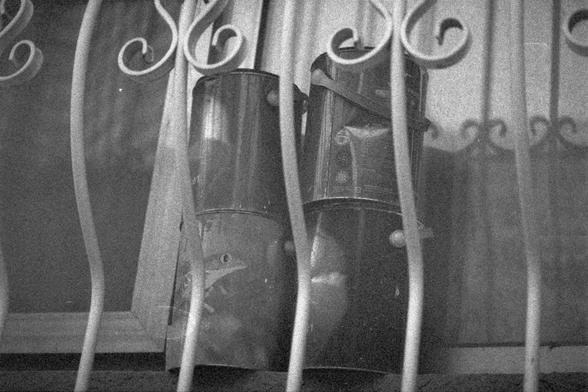 Ilford HP5 Plus 400 (FF)

English Alt Text: Four cylindrical paint buckets sit behind an ornate metal grille, possibly on a window ledge. The buckets have visible markings, suggesting industrial use. The black-and-white photo has a grainy texture, enhancing its nostalgic feel. The grille features curved, decorative patterns, contrasting with the utilitarian devices. The image blends industrial history with artistic architecture.

中文替代文本： 四只圆柱形油漆桶放置在一个装饰性金属栅栏后面，可能位于窗台上。桶上有清晰标记，显示其工业用途。黑白照片呈颗粒质感，…