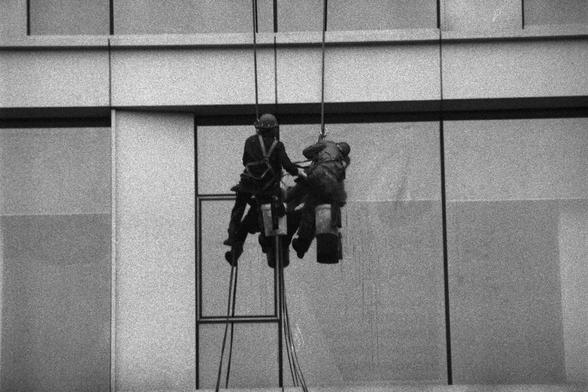 Ilford HP5 Plus 400 (FF)

English Alt Text: Two workers are suspended by ropes, cleaning or maintaining the windows of a modern high-rise building. They wear safety harnesses and carry buckets, emphasizing the risk and skill involved. The building features large glass panels and sleek architectural lines. The image captures teamwork, urban labor, and the importance of safety in high-altitude tasks.

中文替代文本： 两名工人通过绳索悬挂在现代高楼外，进行窗户清洁或维护工作。他们佩戴安全吊带，携带水桶，体现出高空作业的危险性与专业性。建筑外观为大面积玻璃与流线型设计。画面展现了团队协作、城市…