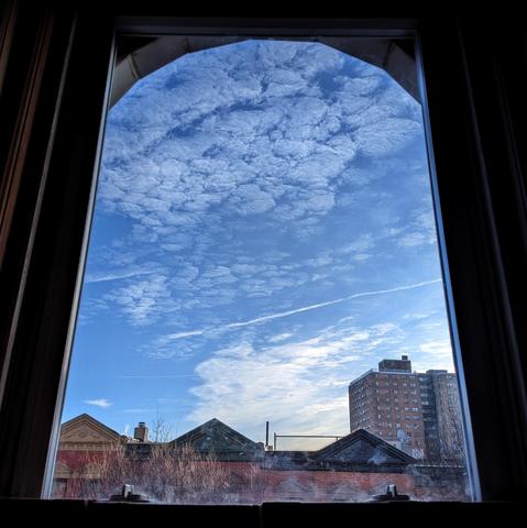 Looking through an arched window ninety minutes after sunrise the blue sky is filled with translucent, puffy white clouds, and a jet trail cuts across the middle sky. Pointed roofs of Harlem brownstones with red brickwork are across the street, and a taller apartment building can be seen in the distance. The leafless tops of two trees are on the bottom and right. The window is grimy on the bottom.