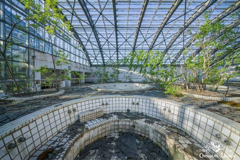 Abandoned indoor spa with broken tiled pools and overgrown vegetation under a large glass ceiling structure.
