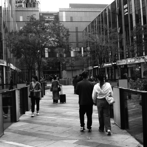 Lucky SHD 400 (6x6)

English Alt Text: A black-and-white photo of a modern city street. Several people walk along a wide pedestrian path lined with trees and tall buildings. The buildings have glass facades and display signs in Chinese characters, including a spa and various shops. Some people carry shopping bags or pull suitcases. The atmosphere is busy but orderly, capturing a typical day in a commercial district. The contrast between the sleek architecture and natural elements like trees add…