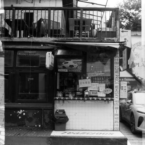 Lucky SHD 400 (6x6)

English Alt Text:
Black-and-white photo of a small urban shop. The storefront is cluttered with signs in Chinese and English. A TV screen displays a soccer ball and promotional text. Bottles and packages are visible inside, suggesting snacks and beverages. A sign reads "LOW PRICE WHISKY." Above the shop is a balcony with chairs and tables. A car is parked nearby. The ground has the word "Delicious" printed. The scene captures dense commercial activity, layered signage, and …