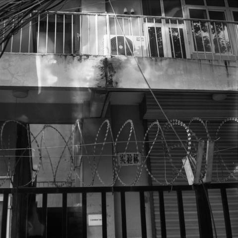 Lucky SHD 400 (6x6)

English Alt Text:
Black-and-white image of a deteriorating urban building. A balcony with barred windows and an air conditioning unit is visible. The exterior wall is stained and damaged. In the foreground, a metal fence topped with coiled barbed wire suggests restricted access. Electrical wires hang across the top of the frame. A sign with Chinese characters ("实验楼") is mounted on the wall behind the fence, partially obscured by the wire. The gritty textures and stark light…