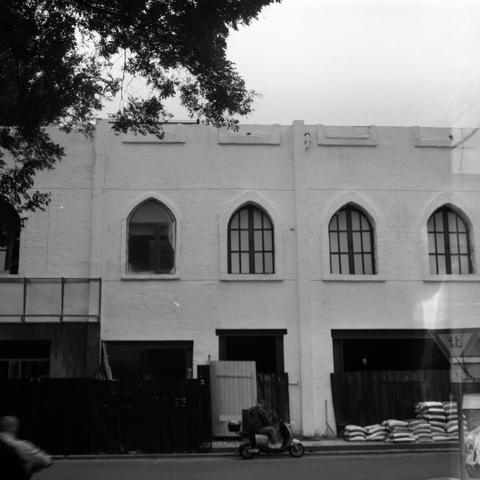 Lucky SHD 400 (6x6)

English Alt Text:
Black-and-white photo of a two-story building. The upper floor features five tall, arched windows with decorative muntins, evoking Gothic architectural style. The textured exterior wall adds visual depth. The lower floor is under renovation, with open doorways, wooden fencing, and construction materials like stacked bags and planks. A person rides a scooter in the foreground, while another figure is partially visible on the left. Tree branches extend into …
