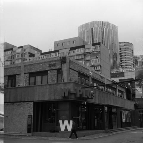 Lucky SHD 400 (6x6)

English Alt Text:
Black-and-white cityscape showing architectural contrast. In the foreground, a modern commercial building labeled "W-PLUS" with a large white "W" at the entrance. Its brick facade and glass windows suggest contemporary design. A person walks past the entrance. Above and behind are mid-rise residential buildings with balconies and air conditioning units. In the background, sleek high-rise towers with curved glass exteriors dominate the skyline. The image ca…