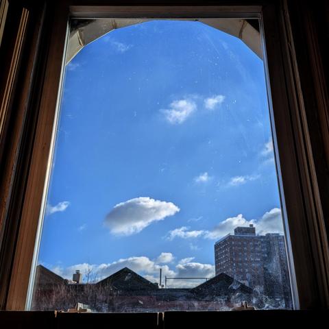 Looking through an arched window three hours after sunrise the bright blue sky is marked with puffy white clouds. Pointed roofs of Harlem brownstones are silhouetted across the street, and a taller apartment building can be seen in the distance. The window is grimy on the bottom.