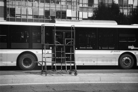 ERA 100 (FF)

English Alt Text:
A black-and-white photo of a city bus parked on a street. In front of the bus is a metal rolling staircase or platform with handrails, likely used for maintenance or boarding. The bus has Chinese characters "北京公交" (Beijing Public Transport) and the number "263400" printed on its side. The background features a modern glass building with reflections. The composition highlights urban infrastructure and public transportation.
中文替代文字：
这是一张黑白照片，展示了一辆停在街边的城市公交车。公交车前方有一…