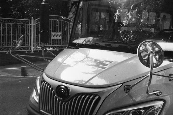 ERA 100 (FF)

English Alt Text:
A black-and-white photo of the front of a school bus. The bus has a large, rectangular grille with a circular emblem at its center. Two side mirrors extend outward from the front, and the windshield reflects trees and surroundings. A bold number "4" is displayed in the top left corner of the windshield, possibly indicating the bus route. The hood features partially visible stylized text or graphics. Behind the bus is a gated fence with a sign that includes the le…