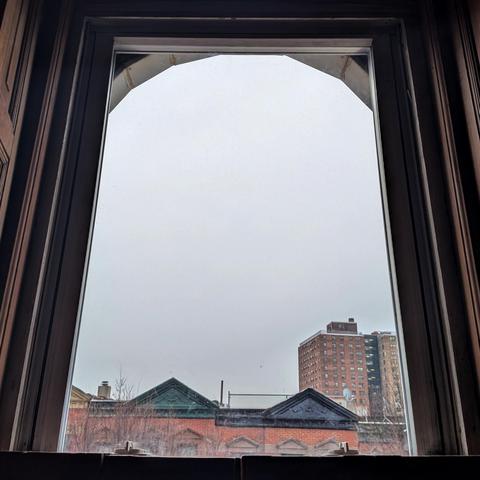 Looking through an arched window an hour after sunrise the sky is a flat gray mass. Pointed roofs of Harlem brownstones with red brickwork are across the street, and a taller apartment building can be seen in the distance. The leafless tops of two trees are on the bottom and right.