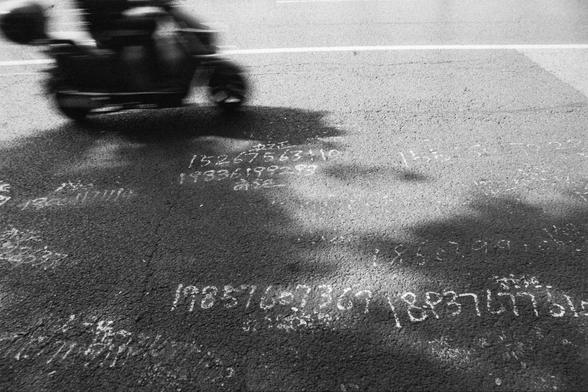 ERA 100 (FF) 

English Alt Text:
A black-and-white photo captures a person riding a scooter on a paved urban road. The motion blur suggests speed and movement. The pavement is covered with handwritten phone numbers and text in white chalk, likely informal advertisements. Numbers such as “15267563110” and “19838199289” are visible. Shadows from nearby trees stretch across the road, adding texture and contrast. The scene evokes a gritty, everyday city moment with a mix of motion and static detail…