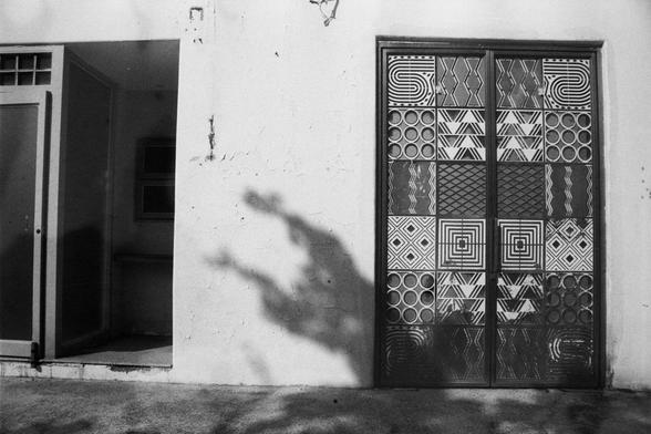 ERA 100 (FF)

English Alt Text:
A black-and-white image of a textured wall with two distinct sections. On the left is a modern doorway leading into a room with a window. On the right is a decorative double door featuring geometric patterns like spirals, diamonds, and zigzags. The wall shows signs of wear with cracks and peeling paint. Tree shadows fall across the surface, adding depth. The juxtaposition of plain and ornate elements creates visual intrigue.
中文替代文本：
这是一张黑白照片，展示一面有质感的墙壁，分为两个部分。左侧是…
