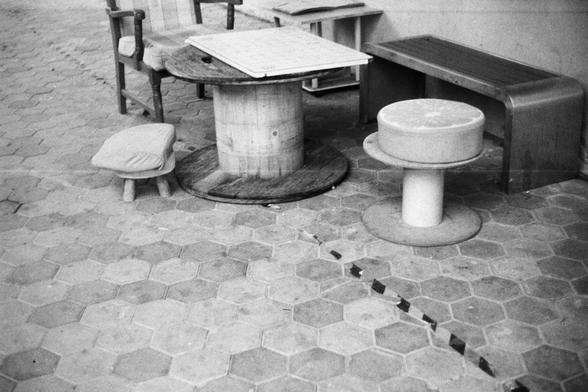 ERA 100 (FF)
English:
This black-and-white image shows an outdoor area with hexagonal patterned tiles on the ground. In the center, there is a round wooden table with a white tabletop, surrounded by various seating options: a wooden stool with a cushion, a round stool made from a cable spool, and a bench. The scene gives a rustic, creative vibe, as if it is a DIY outdoor seating area.
Chinese:
这张黑白照片展示了一个户外区域，地面铺着六边形图案的瓷砖。中央有一张圆形的木桌，桌面是白色的，周围摆放着不同的座位：一个带垫子的木凳、一个由电缆卷筒改造的圆凳，以及一个长凳。整个场景散发出乡村和创意的气息…