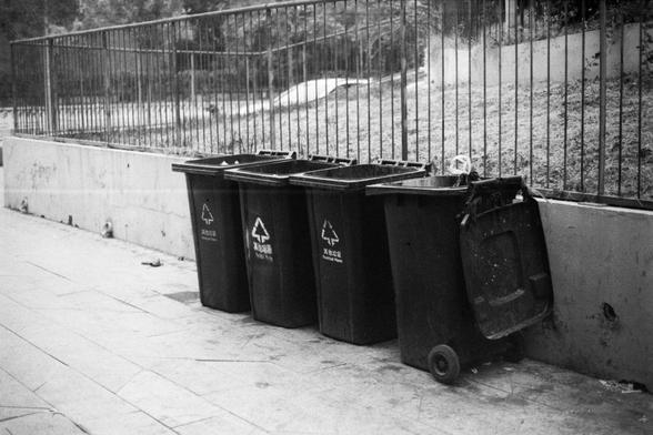 ERA 100 (FF)
English:
This black-and-white image shows a row of four black recycling bins lined up against a low wall and a metal fence. Each bin has a recycling symbol and Chinese text indicating they are for recyclable waste. The bins are placed on a paved sidewalk, with a grassy hill and more fencing in the background, suggesting a public or park area.
Chinese:
这张黑白照片展示了四个黑色的回收垃圾桶，整齐地排列在一面矮墙和金属栅栏旁。每个垃圾桶上都有回收标志和“可回收物”的中文字样。垃圾桶放在铺砌的人行道上，背景是草地和更多的栅栏，暗示这是一个公共区域或公园。