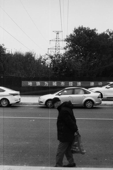 ERA 100 (FF)
English:
This black-and-white photo captures a street scene. A person is walking on the sidewalk, carrying a bag. In the background, there are cars parked along the road, and a large billboard with Chinese characters that reads "地铁为城市提速建设" (Subway accelerates city construction). There is also a power transmission tower and trees lining the street, adding depth to the urban landscape.
Chinese:
这张黑白照片展示了一个街道场景。一个人正在人行道上行走，手里提着一个袋子。背景中停着几辆汽车，还有一个大型广告牌，上面写着“地铁为城市提速建设”的字样。远处有输电塔和排列整齐的树木…