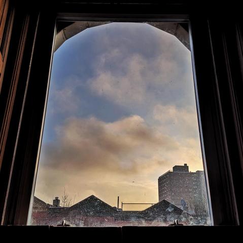 Looking through an arched window an hour after sunrise the blue sky is filled with diffuse white and yellow clouds. Pointed roofs of Harlem brownstones are silhouetted across the street, and a taller apartment building can be seen in the distance. The window is grimy on the bottom.