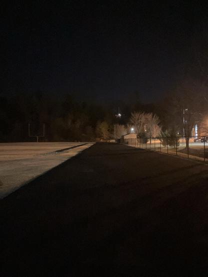 Looking down the main straightaway at night towards all the pine trees at the far end. Also visible in the far distance are the yellow uprights of an American football goalpost. 