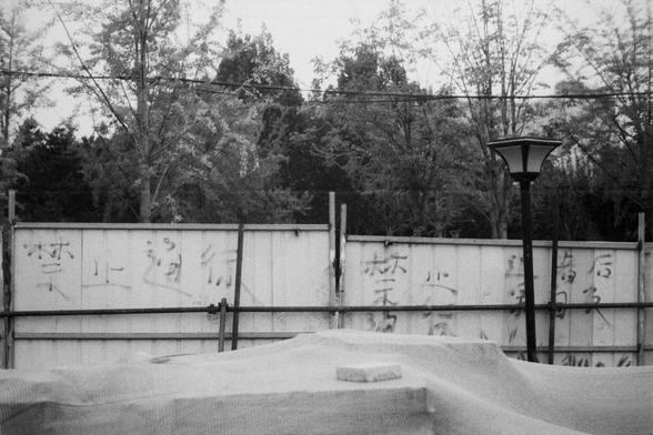 ERA 100 (FF)

English Alt Text:
A monochrome image of a fenced-off area, likely under construction or restricted. The metal fence bears handwritten Chinese characters: “No entry” and “No peddling.” Behind the fence are tall trees and a power line running horizontally. In the foreground, a covered object with a brick on top sits near a streetlamp. The scene feels quiet and controlled, with signs of urban regulation and exclusion.
中文替代文字：  
这是一张黑白照片，显示一个被金属围栏封锁的区域，可能是施工或限制通行的场所。围栏上手写着中文：“禁止通行”和“禁…