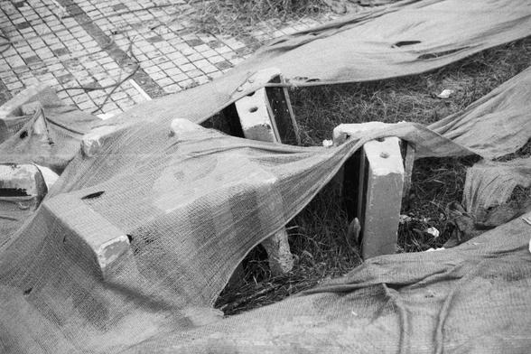 ERA 100 (FF)

English Alt Text:
A grayscale photo showing several concrete blocks arranged on a grassy patch. Some blocks stand upright, others lie flat. They are partially covered with coarse, weathered fabric—possibly burlap—with visible holes and frayed edges. The background includes tiled pavement and more grass. The scene suggests a temporary construction setup or abandoned materials, with textures of rough stone and worn cloth.
中文替代文字：  
这是一张灰度照片，画面中有几块混凝土砖块摆放在草地上，有的竖立，有的平躺。砖块部分被粗糙、风化的布料覆…
