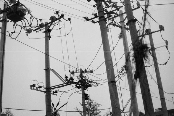 ERA 100 (FF)
English Alt Text:
A black-and-white image capturing a dense network of utility poles and electrical wires. Multiple concrete poles are fitted with insulators, transformers, and other components. Wires crisscross in various directions, forming a complex web. The monochrome palette emphasizes the geometry and industrial feel of the infrastructure. The image reflects the intricacy and importance of urban power distribution systems.
中文替代文字：  
这是一张黑白照片，展示了密集的电力基础设施网络。多个混凝土电线杆上安装有绝缘器、变压器…