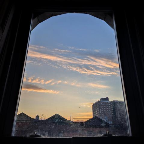 Looking through an arched window fourteen minutes after sunrise the blue sky is marked with horizontal streaks of white clouds which are underlit orange by the rising sun. Pointed roofs of Harlem brownstones are silhouetted across the street, and a taller apartment building can be seen in the distance. The window is grimy on the bottom.