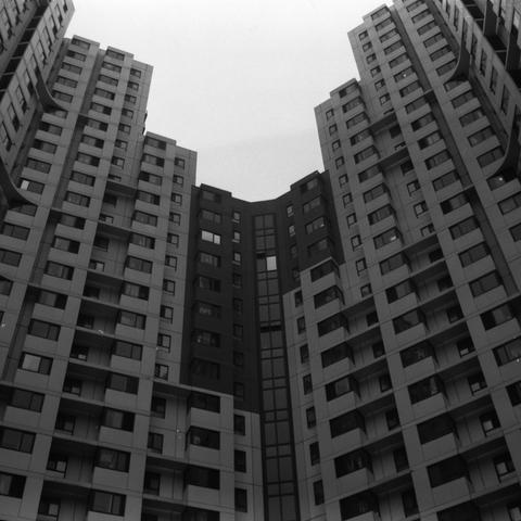 Lucky SHD 400 (6x6)

English Alt Text:
A dramatic black-and-white photo of tall modern apartment buildings forming a semi-enclosed courtyard. The buildings feature repetitive patterns of windows and balconies, with vertical and horizontal lines creating a geometric rhythm. The central building has a darker facade with a vertical strip of windows, possibly indicating a stairwell. The photo is taken from a low angle, emphasizing the towering height and density of the structures. The monochrome pa…
