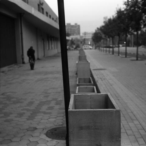 Lucky SHD 400 (6x6)

English Alt Text:
A quiet urban street scene in black and white. A row of large, empty concrete planters lines the sidewalk, creating a strong sense of depth and perspective. On the left, a building with multiple windows stretches into the distance, and a lone person walks away from the camera. On the right, evenly spaced trees add contrast to the geometric shapes of the planters. The street is calm, with no visible traffic, and the composition emphasizes symmetry and solit…