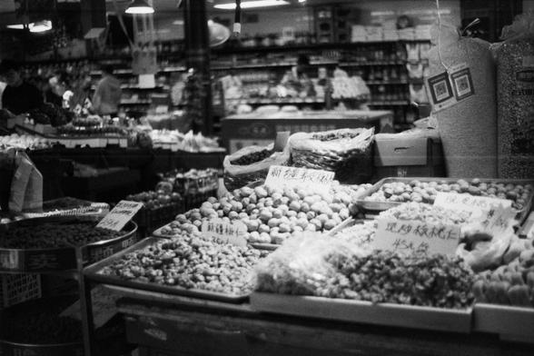 ERA 100 (FF)

English Alt Text: Black-and-white image of a market section selling dried goods and nuts. Trays and containers hold various items like walnuts and almonds. Handwritten Chinese signs indicate names and prices, such as “Xinjiang walnut kernels 25 yuan per jin,” “45 yuan per jin,” “52 yuan per jin,” and “Badam nuts.” Shelves in the background are stocked with packaged goods. A few shoppers are visible. The image evokes a nostalgic feel, highlighting the tactile and sensory richness o…