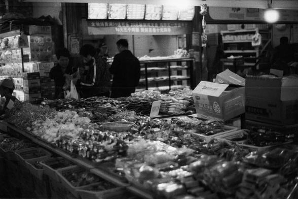ERA 100 (FF)

English Alt Text: Black-and-white photo of a busy indoor market. Tables and shelves are filled with packaged snacks and dried goods in baskets and boxes. Shoppers browse and interact with products. The lighting is dim, giving a vintage feel. Chinese signs above the counter advertise promotions: “Spend 33 yuan to receive a free gift” and “Three items for 1 yuan, limited to first ten daily.” A box in the foreground displays the brand “Orion” and “Orion Pie.” The scene captures the l…