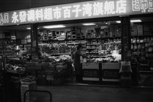 ERA 100 (FF)

English Alt Text: Color image of a Chinese supermarket entrance. A shopper is visible inside, browsing shelves filled with packaged foods, fresh produce, and bottled items. The store is brightly lit and well-stocked. Above the entrance is a glowing sign in Chinese: “Entrance Yongfa Seasoning Supermarket Baiziwan Flagship Store.” Promotional signs on boxes in the foreground read “PROMOTION” in both Chinese and English. The image reflects a modern urban shopping experience in China,…