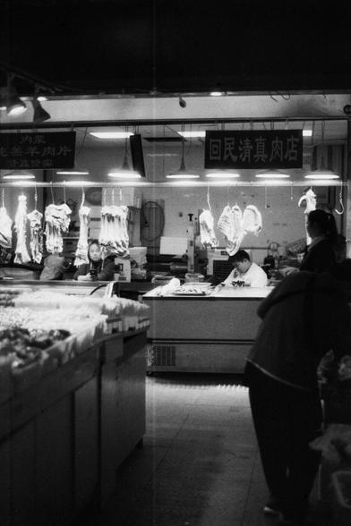 ERA 100 (FF)

English Alt Text: Black-and-white photo of a traditional meat market. Several large cuts of meat hang from metal hooks under bright lights. Workers behind the counter are preparing meat, one actively slicing. The market is busy and utilitarian, with counters displaying various meat products. Two signs in Chinese are visible, indicating halal offerings for the Hui Muslim community. The scene captures a culturally rich, bustling environment focused on meat processing and sales.

中文替…