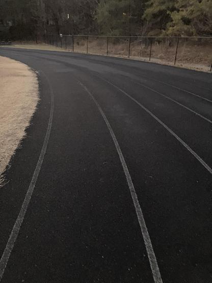 The lanes of the track curve to the right towards the backstretch. There is a fence with woods beyond. Inside the track is a grass practice field, the warm weather grasses a dormant tan color. 