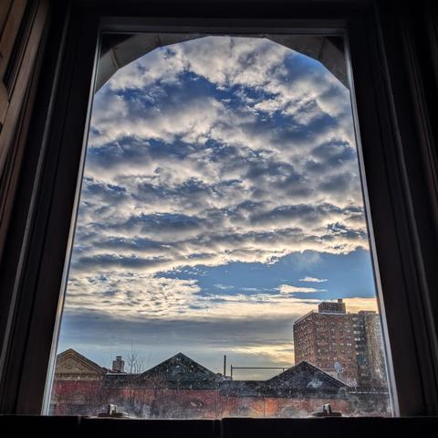 Looking through an arched window forty-eight minutes after sunrise the blue sky is filled with complex, chunky white clouds. Pointed roofs of Harlem brownstones with red brickwork are across the street, and a taller apartment building can be seen in the distance. The window is grimy on the bottom.