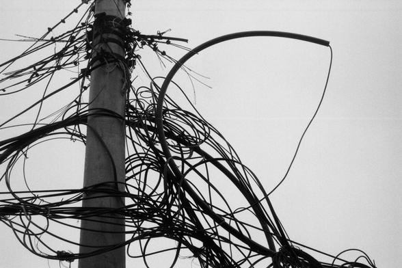 ERA 100 (FF)

English Alt Text:
A black and white image of a utility pole overwhelmed by a chaotic tangle of wires. The wires loop, twist, and hang in various directions, some appearing disconnected or frayed. The dense mass of cables creates a visual maze, suggesting poor maintenance or infrastructure overload. The pole itself is weathered, and the scene hints at potential hazards. The image emphasizes the complexity and disorder of urban electrical systems.
中文替代文字：
这是一张黑白照片，展示了一根电线杆被大量杂乱电线缠绕的…