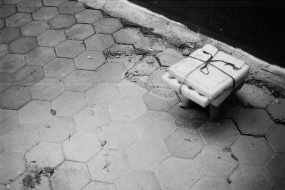 ERA 100 (FF)

English Alt Text:
A black and white photo of an outdoor tiled surface made of hexagonal tiles. The tiles are worn and uneven near the top edge, with some broken pieces and scattered debris. In the upper right corner, a small stack of square slabs tied with string sits atop a short stand or stool. The slabs appear heavy and utilitarian, possibly construction materials. The scene captures a gritty urban texture, with strong contrasts between the geometric tile pattern and the roughn…
